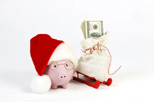 Tirelire rose avec chapeau et lunettes Santas debout à côté du traîneau rouge avec sac Santas et pile d'argent billets de cent dollars américains sur la neige — Photo