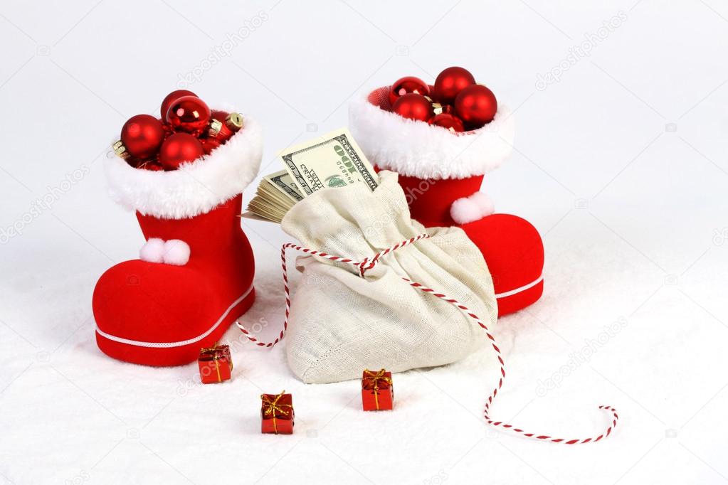 Two Santas boots with red mat christmas balls and Santas bag with stack of money american hundred dollar bills and three gifts with gold bow on snow
