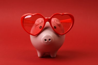 Piggy bank in love with red heart sunglasses standing on red background clipart