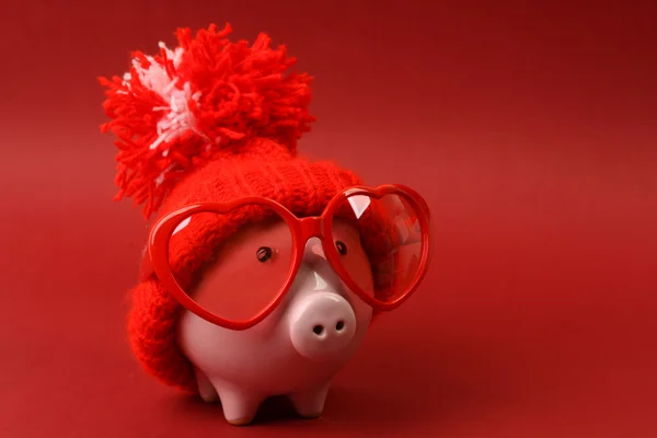 Piggy bank in love with red heart sunglasses with red hat and pom-pom standing on red background — Stock Photo, Image