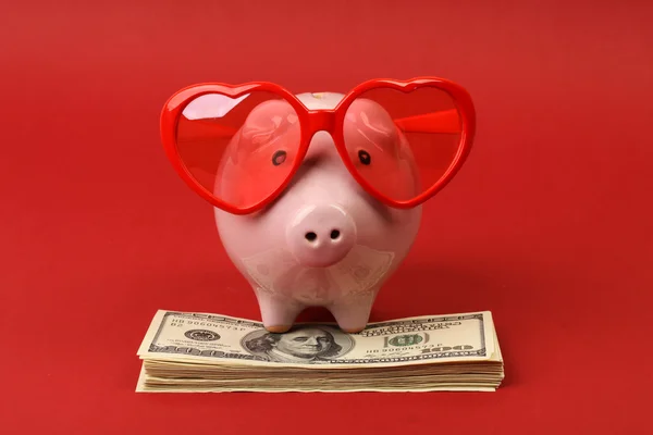 Hucha en el amor con gafas de sol corazón rojo de pie en la pila de billetes de cien dólares americanos en el fondo rojo — Foto de Stock