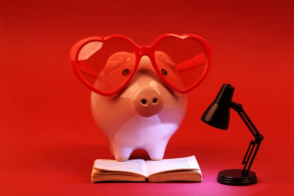 Piggy banco no amor com óculos de sol coração vermelho lendo um livro e brilhante lâmpada preta no fundo vermelho — Fotografia de Stock