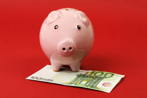 Pequeña alcancía rosa de pie en billetes en euros hunderd sobre fondo rojo —  Fotos de Stock