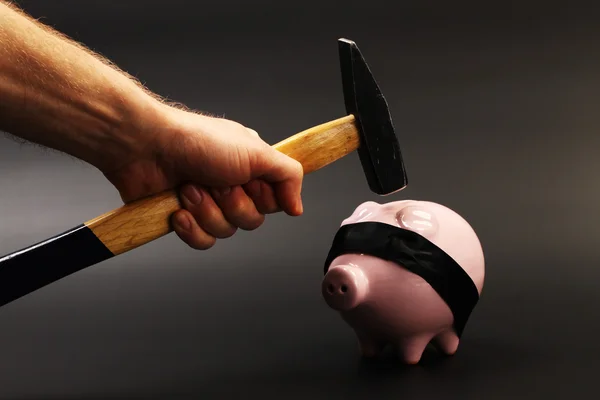 Uma mão segurando um martelo que é levantado acima de um porquinho rosa de cabeça para baixo com venda preta em pé sobre fundo preto — Fotografia de Stock