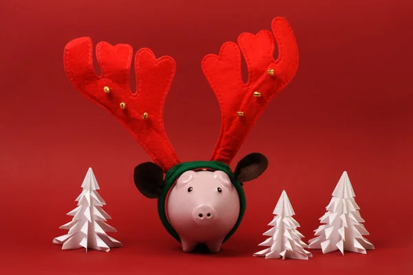 Piggy bank with reindeer horn and jingle bells standing on red background with three white origami trees Christmas background — Stock Photo, Image