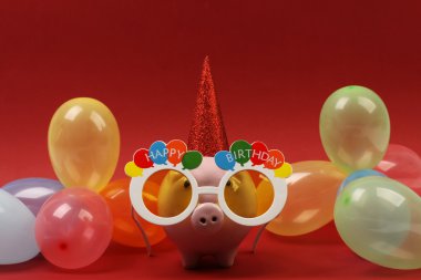 Güneş gözlüğü mutlu doğum günü, parti şapkası ve kırmızı bir arka plan üzerinde renkli Parti balonları ile Piggy banka