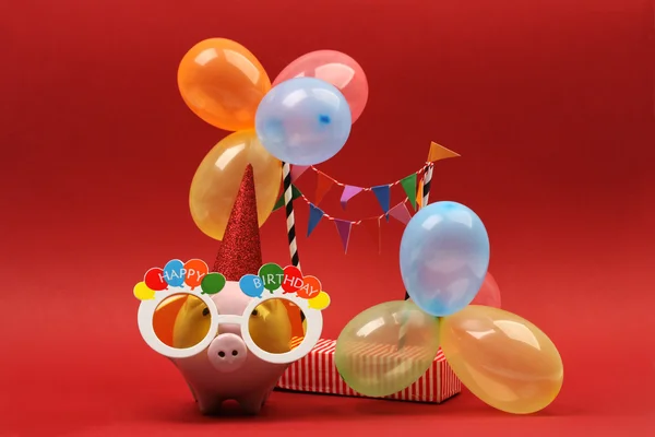 Piggy bank com óculos de sol Feliz aniversário, chapéu de festa e balões multicoloridos no fundo vermelho — Fotografia de Stock