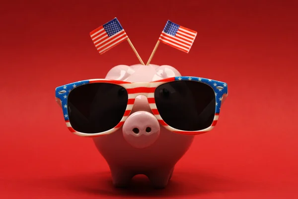 Piggy bank com óculos de sol retro com bandeira dos EUA e duas pequenas bandeiras dos EUA no fundo vermelho — Fotografia de Stock