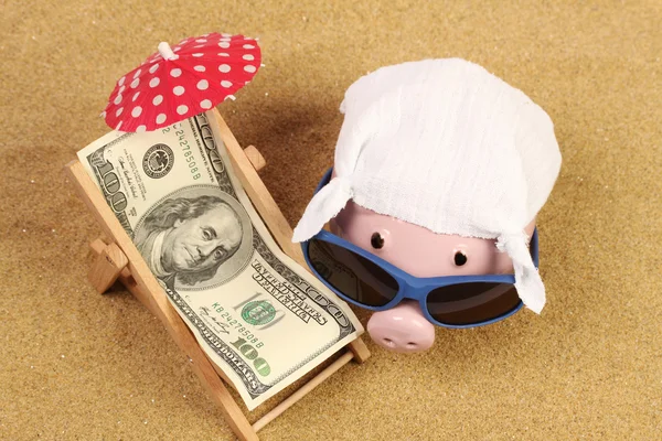 Summer Piggy Bank com óculos de sol e lenço ao lado da cadeira de praia com toalha de greenback cem dólares e guarda-sol vermelho na praia de areia — Fotografia de Stock