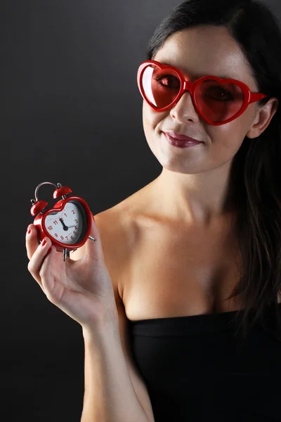 Young and pretty woman with heart shaped glasses and heart-shape alarm clocker on black background — Stock Photo, Image