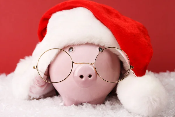 Hucha rosa con sombrero de Santa Claus con pompón y gafas de pie sobre nieve blanca sobre fondo rojo —  Fotos de Stock