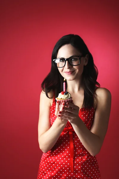 Jolie fille caucasienne avec des lunettes soufflant bougie sur son gâteau tasse sur fond rouge Images De Stock Libres De Droits