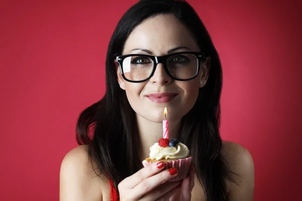 Jolie fille caucasienne avec des lunettes soufflant bougie sur son gâteau tasse sur fond rouge Images De Stock Libres De Droits