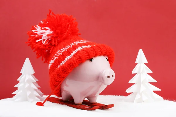 Piggy box with red hat with pompom standing on red ski and ski sticks on snow and around are snowbound trees on red background — Stock Photo, Image