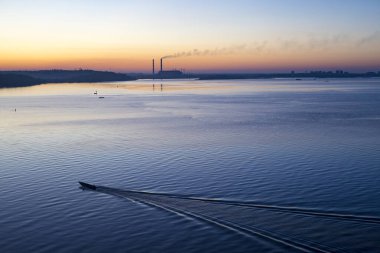 Gün batımında şehrin arka planında, nehirde tekne. Büyük bir nehir ve tekneyle aydınlık bir yaz günbatımı.