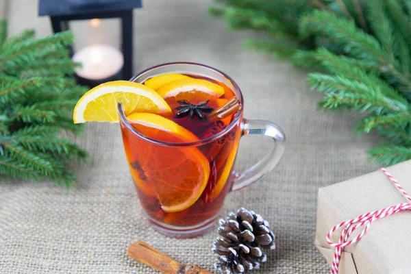 A transparent cup with hot mulled wine with oranges and a cinnamon stick on a beige background, near green spruce branches. New Year\'s drink, hot Christmas wine with spices.