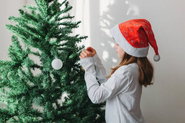 Açık renk saçlı bir kız Noel Baba şapkasıyla Noel ağacını Noel süsleriyle süslüyor. Yeni yıl, geleneksel aşçılık kutlaması..