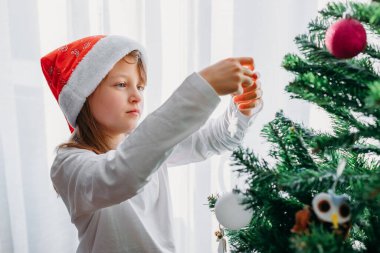 Noel Baba şapkalı tatlı, koyu renk saçlı bir kız ağacı Noel oyuncaklarıyla süslüyor. Yeni yıl ve Noel için dekorasyon. Dekorasyon