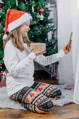 Çevrimiçi Yeni Yıl ve Noel tebrikleri. Sevimli gülen kız internetteki görüntülü konuşma için cep telefonu kullanıyor. Aileleri, aileleri ya da arkadaşlarını arıyor. Arka planda Noel ağacı.