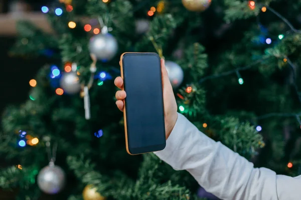 Mobile phone in the hands of a girl against the background of a New Year tree. Online greetings for the new year. Online shopping.