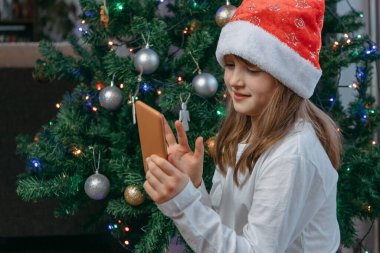 Çevrimiçi Yeni Yıl ve Noel tebrikleri. Sevimli bir kız internette görüntülü konuşma için cep telefonu kullanıyor. Aileleri, aileleri ya da arkadaşlarını arıyor..