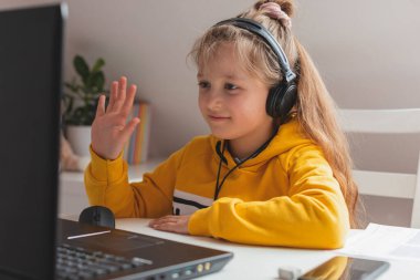 Çevrimiçi çalışma, evde uzaktan öğrenme. Kulaklıklı sevimli sarışın beyaz kız bilgisayar ya da dijital tablet üzerinde ödev yapıyor. Video konferansı ve arkadaşlarla çevrimiçi toplantı.
