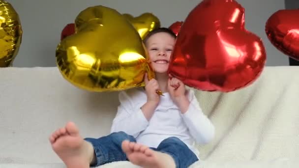 情人节视频中，可爱的男孩沉溺于一个红色和金色的心形气球在一个轻便的沙发上。快乐，快乐，爱 — 图库视频影像