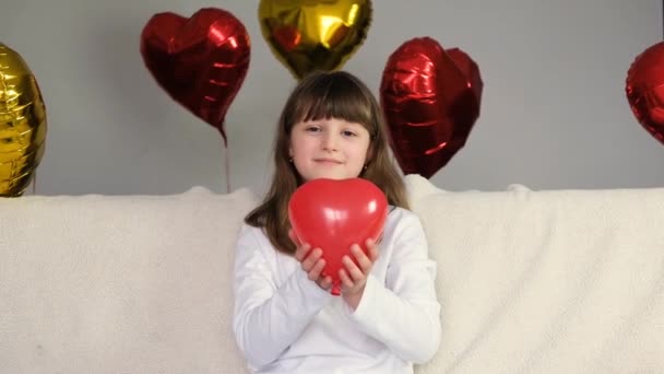 Valentinstag. Ein süßes Mädchen auf dem Sofa, umgeben von Luftballons, hält einen roten herzförmigen Luftballon in den Armen. Zeitlupe — Stockvideo