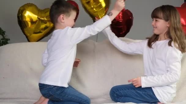 Lindo, lindo chico y chica se están divirtiendo con globos de corazón en un sofá beige claro. Día de San Valentín, Día de San Valentín. Movimiento lento — Vídeo de stock