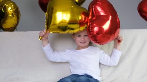 Dia dos Namorados vídeo, menino bonito está se entregando com um balão vermelho e dourado em forma de coração em um sofá leve. Diversão, alegria, amor. Movimento lento — Vídeo de Stock