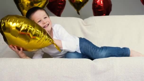 Garoto bonito no sofá está se divertindo e se divertindo com um balão de coração dourado. Feriado Dia dos Namorados. Movimento lento — Vídeo de Stock