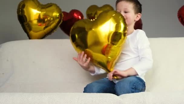 Lindo chico en el sofá es complacer y divertirse con un globo de oro corazón. Día de San Valentín. Movimiento lento. — Vídeos de Stock