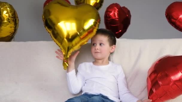 Lindo chico divirtiéndose, complaciendo o jugando en un sofá ligero con globos en forma de corazón. Día de San Valentín. Mociones lentas — Vídeo de stock