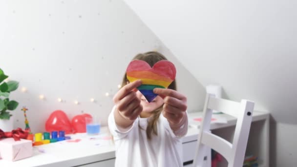 Nahaufnahme eines regenbogenfarbenen Herzens in den Händen eines niedlichen kaukasischen Mädchen.Freie Liebe, lgbt. Symbol der Liebe, Valentinstag, Vater- und Muttertag. Zeitlupe — Stockvideo