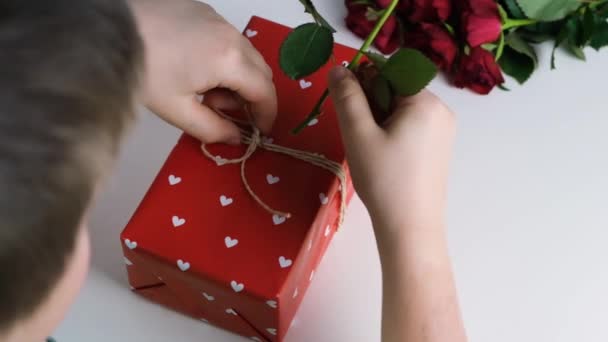 Chłopiec ozdabia pudełko prezentem z czerwoną różą. Dekorowanie i pakowanie prezentów, prezentów wakacyjnych. Zwolnione ruchy — Wideo stockowe