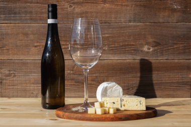 Bir şişe beyaz şarap, bir bardak ve ahşap arka planda parlak yaz güneşinde farklı tipte peynir. Metin için yer