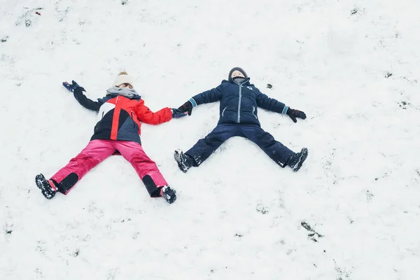 Ragazzo e ragazza, fratello e sorella fanno un angelo di neve nel cortile di una casa nella neve. Giochi per bambini sulla neve, vacanze invernali, giochi e divertimento — Foto Stock