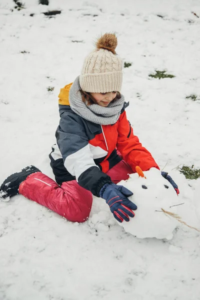 Giochi di neve, intrattenimento e divertimento. Una ragazza caucasica carina scolpisce un pupazzo di neve nel cortile. Decora la testa di un pupazzo di neve. Orientamento verticale, persone autentiche — Foto Stock