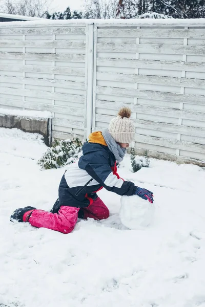 Una ragazza caucasica carina di 9 anni fa rotolare un pupazzo di neve nel cortile di casa. Neve divertimento e giochi, vacanze invernali. Orientamento verticale, Stile di vita. — Foto Stock