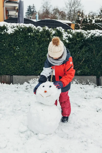 Giochi di neve, intrattenimento e divertimento. Una ragazza caucasica carina scolpisce un pupazzo di neve nel cortile. Orientamento verticale, persone autentiche — Foto Stock