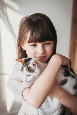 Sekiz yaşında tatlı bir kız, üç renkli kedisine evde sarılıyor. Dikey oryantasyon. Evcil hayvanlara sevgi ve ilgi