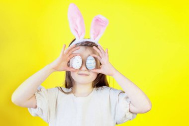 Paskalya konsepti. Tavşan kulaklı şirin bir kızın portresi boyalı yumurtalarla uğraşıyor, sarı arka planda yüzünü örtüyor. Metin için yer