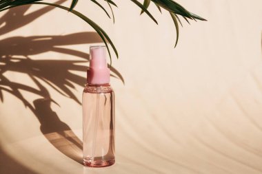 Body care product, lotion, thermal water or liquid oil for moisturizing during the sunny period. Pink jar with liquid on a beige background with the shadow of palm trees. Copy space