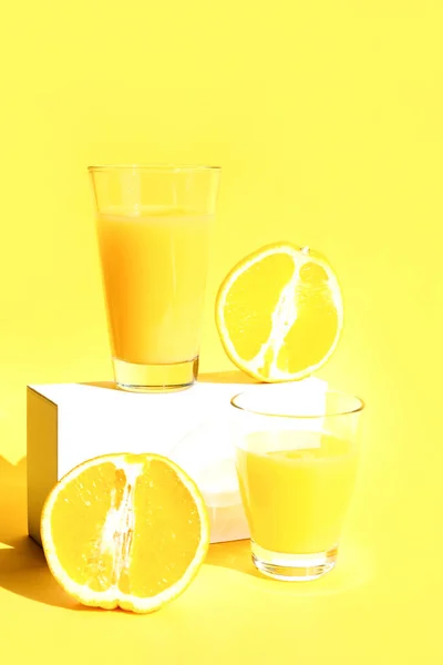 Un bicchiere di succo d'arancia rinfrescante appena spremuto su un piedistallo bianco o un podio con metà arancio. Fondo giallo. Il concetto di uno stile di vita sano, dieta cruda, vegetarianismo. Copia spazio — Foto Stock