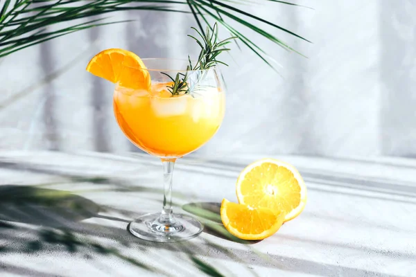 Una copa con un cóctel de verano con una naranja y una ramita de romero bajo una hoja de palma sobre un fondo gris. Lugar para tu mensaje. bebidas frías en el calor —  Fotos de Stock