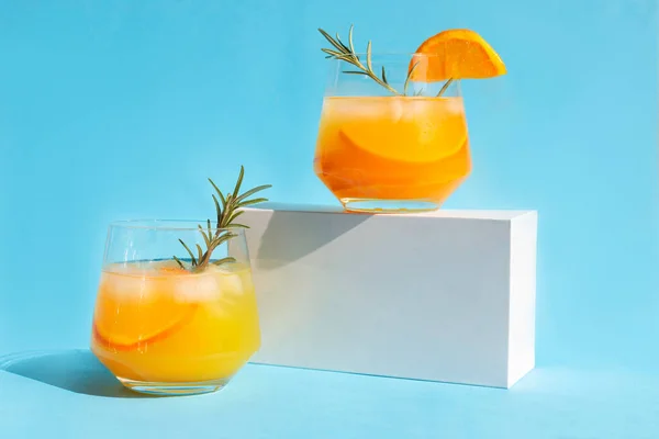 Lemonade, orange cocktail, refreshing summer drink with ice and orange slices in a glass with a sprig of rosemary on a white podium. Trending drinks.