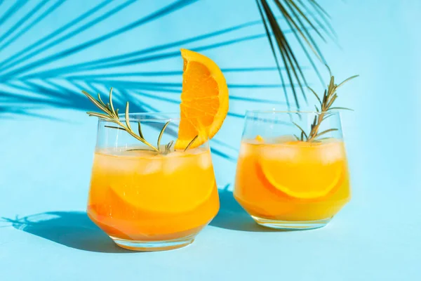 Detailní limonáda, pomerančový koktejl, osvěžující letní nápoj s ledem ve sklenici s rozmarýnem na modrém pozadí pod palmovým listem za jasného slunečného dne. — Stock fotografie