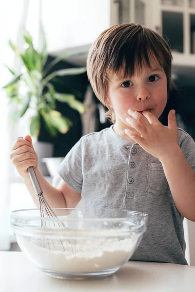 A cute boy, a toddler of Caucasian appearance, cooks in the kitchen and tastes the dough. Vertical photo. Ideal for illustrating real life moments — Photo
