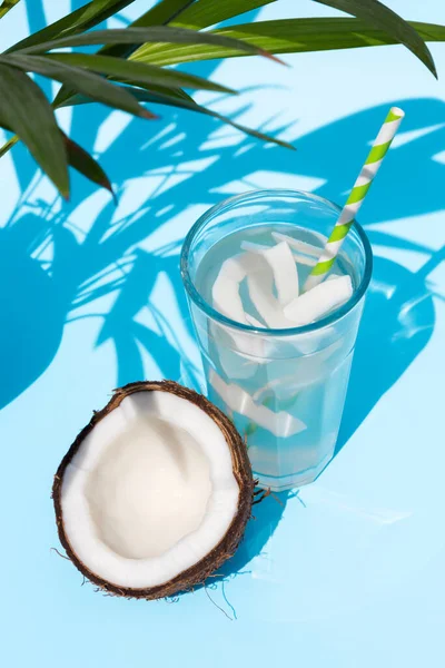 Woda kokosowa w szklance z rurką koktajlową i pół świeżego kokosa w pobliżu na niebieskim tle pod liśćmi palmy w słoneczny dzień. Idealny sok odtruwający — Zdjęcie stockowe