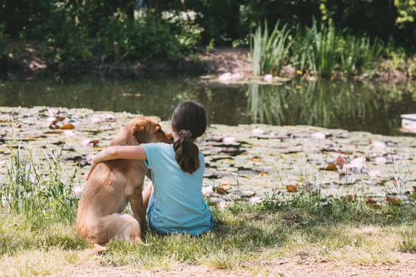 Přátelství zvířat a dětí, lidí. Běloška sedí na břehu rybníka, řeka a objímá labradorské štěně. Letní venkovní rekreace — Stock fotografie